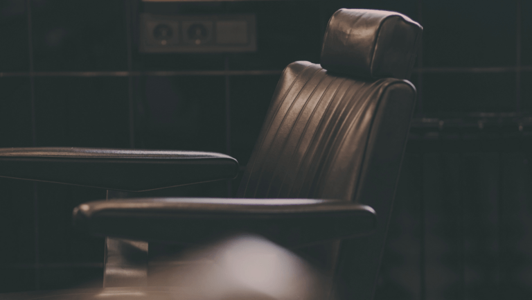 A black barber chair with a chrome base sits in a dark room. The room is so dark that the only thing visible in the image is the barber chair.