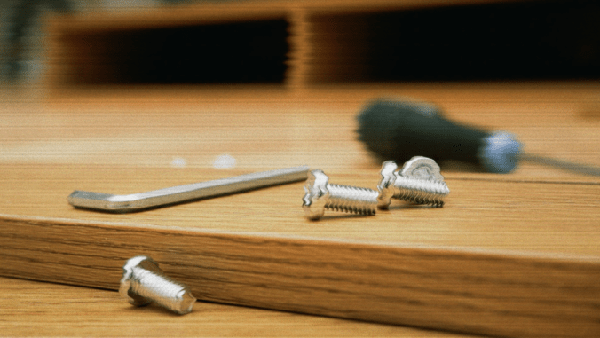 Close-up of an Allen wrench, screws, and a bolt on a wooden surface. A blurred screwdriver is in the background. The image conveys the theme of assembling or repairing furniture or other items.