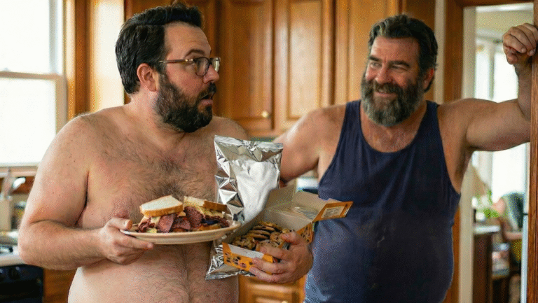 Two bearded men in a kitchen, one shirtless holding a plate of sandwiches with a surprised look, the other in a dark tank top smiling and holding a bag of chips and a box of cookies.