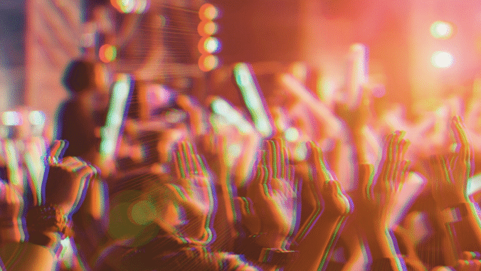 A vibrant crowd with raised hands enjoys a lively concert or festival, illuminated by colorful lights and glowing sticks, creating an energetic and festive atmosphere.