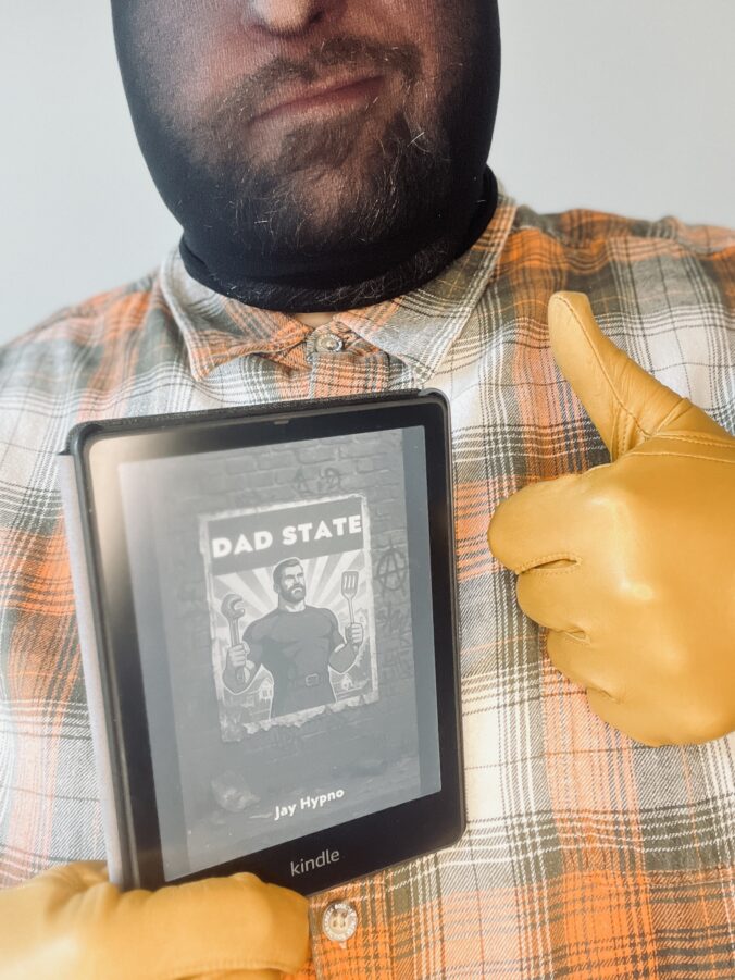 Jay Hypno wearing a plaid shirt and yellow gloves gives a thumbs up while holding a Kindle displaying the cover of the book “Dad State” by Jay Hypno.